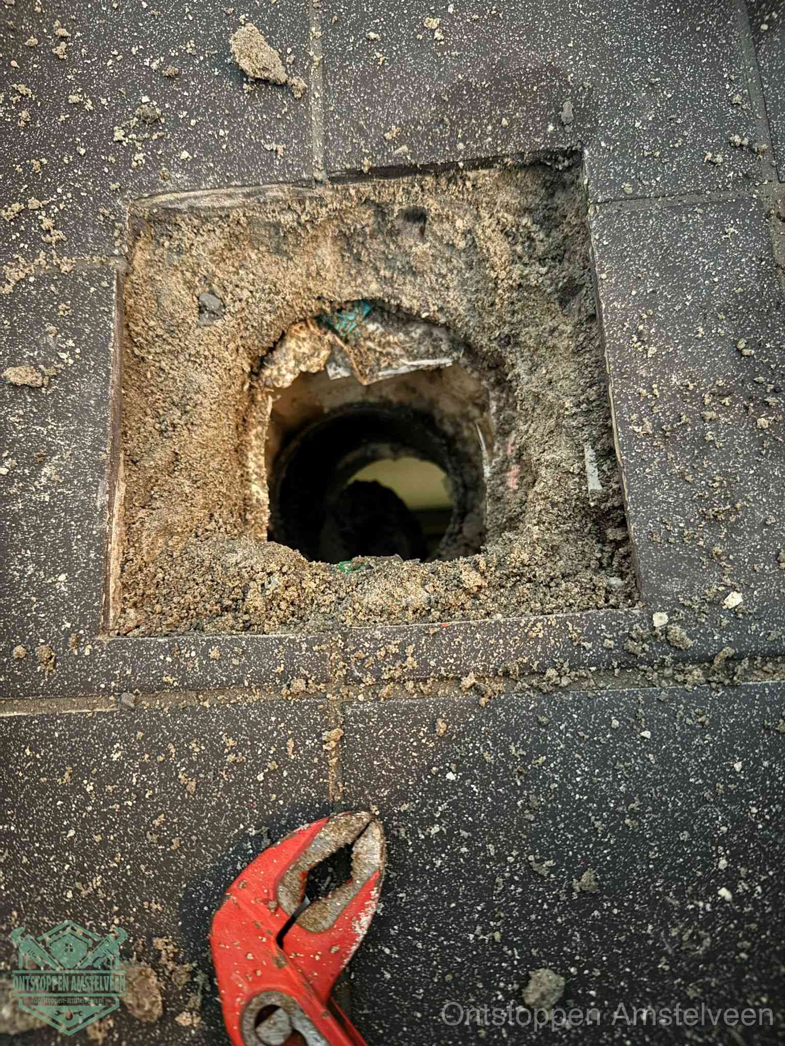 Een geopend rioolputje in een betontegelvloer met losgemaakt rooster en een zichtbare ronde afvoerbuis, het achterhalen waar de afvoerverstopping zit in Amstelveen