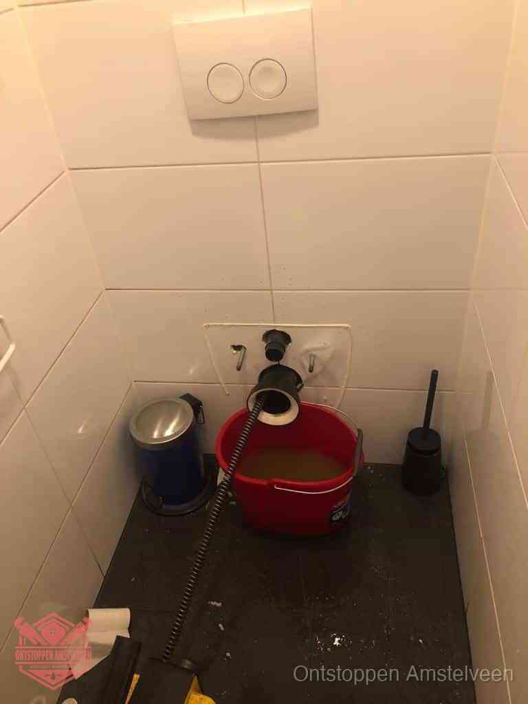 Een ontstopte wand-wc met verwijderde toiletpot waar een ontstoppingsveer in de wanduitgang verdwijnt, het achterhalen waar de verstopping zit in Amstelveen