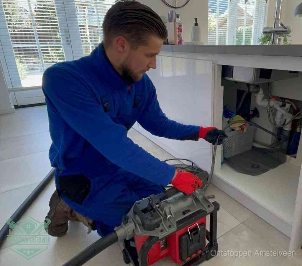 Een monteur met rode handschoenen knielt in een keuken en voert een elektrische ontstoppingsveer in tijdens het professioneel afvoer ontstoppen in Amstelveen