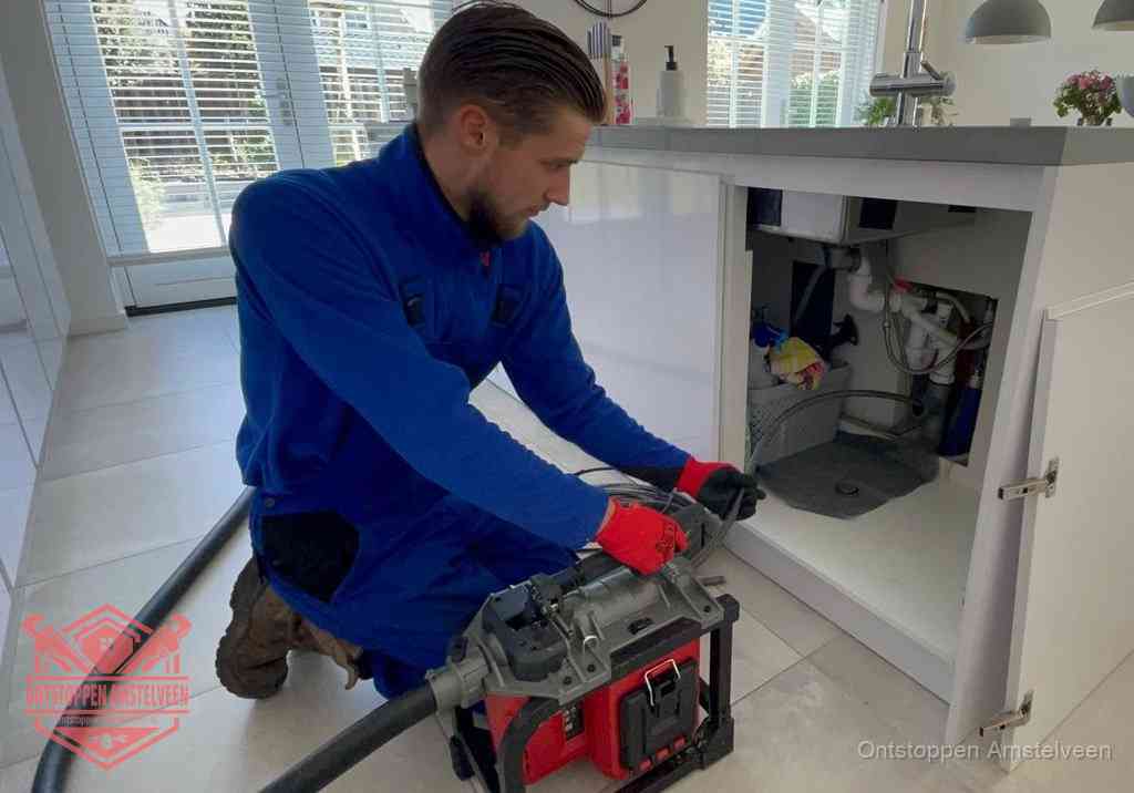 Een monteur voert een professionele elektrische ontstoppingsmachine in de afvoer onder de spoelbak tijdens het gootsteen ontstoppen in Amstelveen